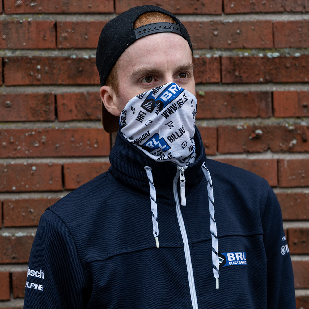 En man med tubhalsduk i vitt, svart och blått mot en tegelvägg.