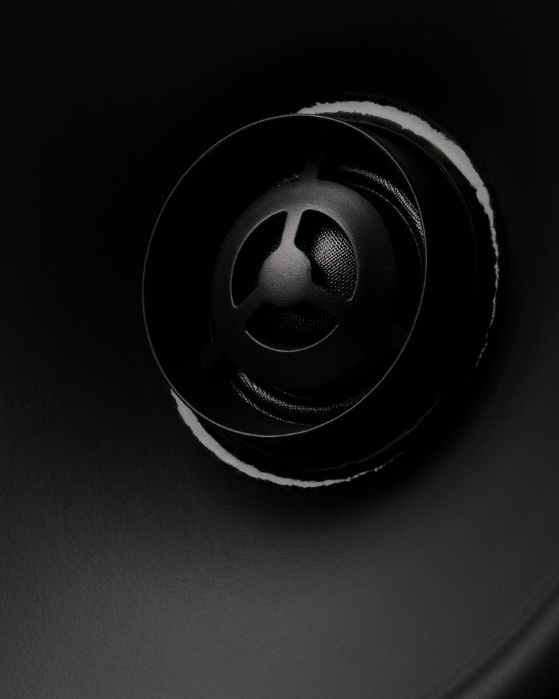 En mattsvart högtalare med ett runt grilleskydd och centralt element.