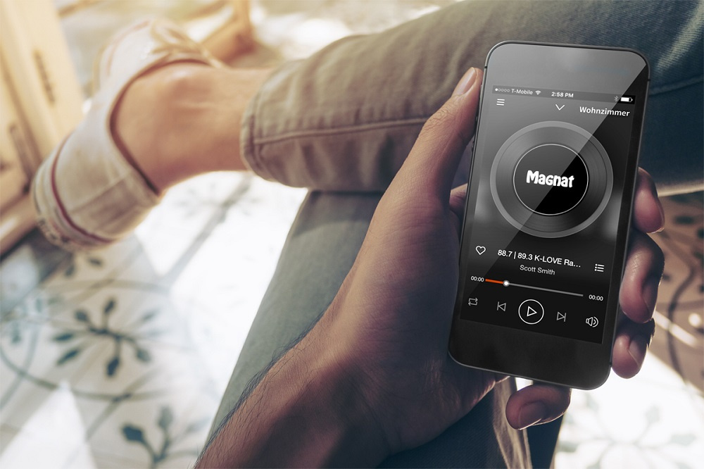 En hand som håller en smartphone med Magnat-app och en avslappnad person i bakgrunden.