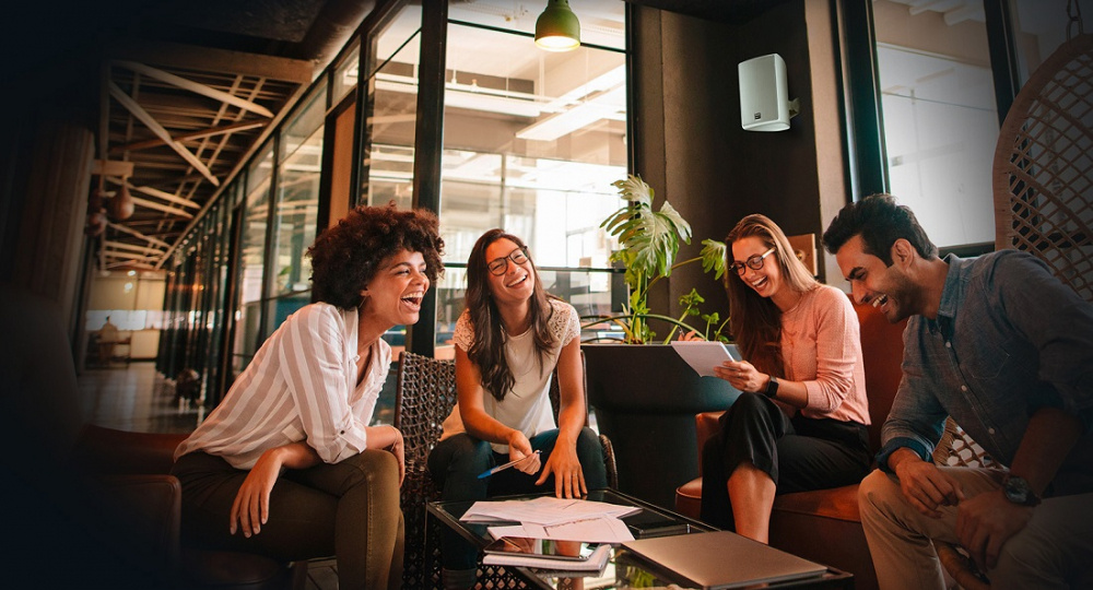 Fyra personer skrattar och pratar i en modern miljö med högtalare på väggen.