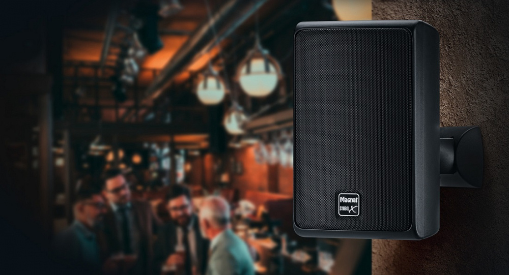 Svart utomhushögtalare monterad på vägg i en baraktig miljö.