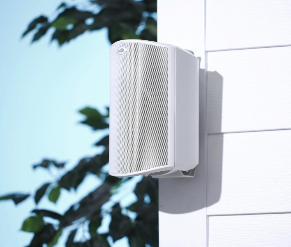 Vita utomhushögtalare monterade på vägg, med grönskande omgivning.