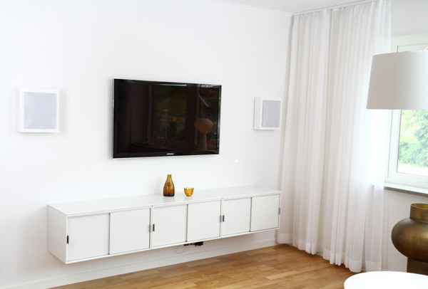 Vitt rum med TV på väggen, låg vit möbel och två vägghögtalare.