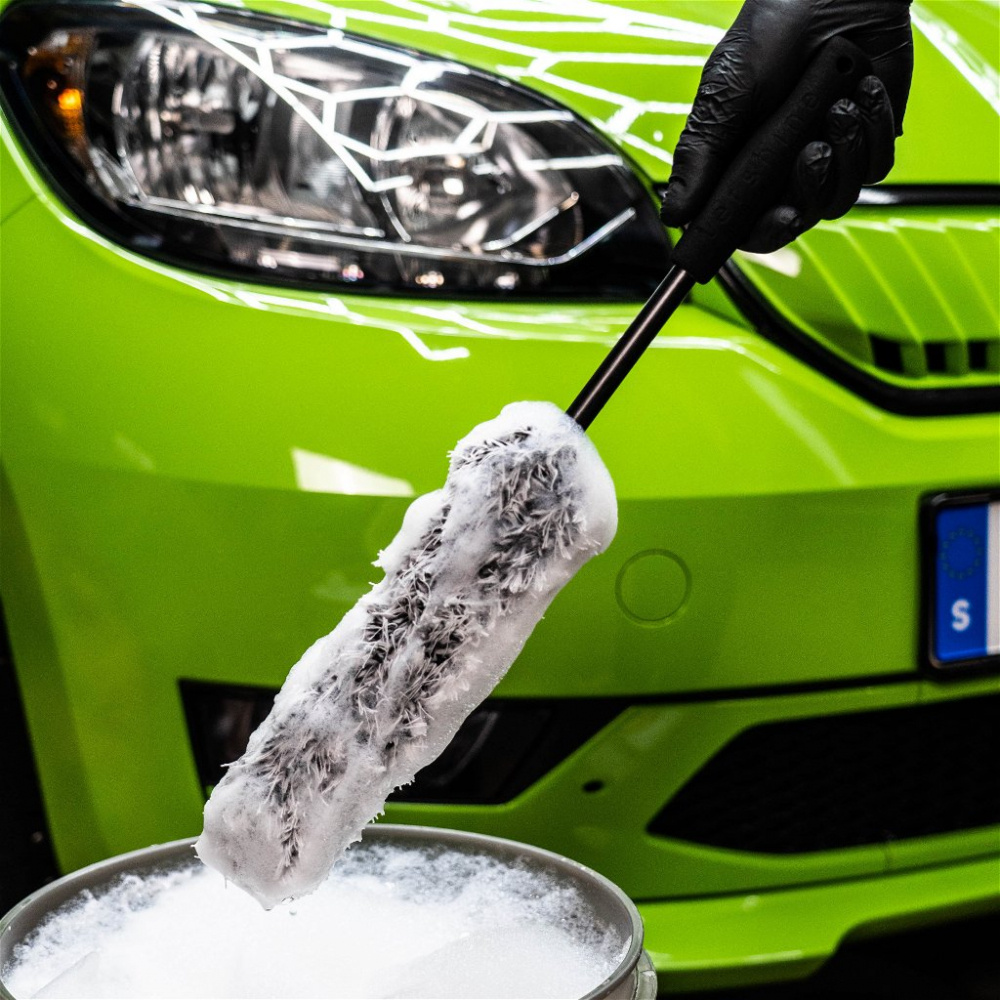 En hand som håller en borst för däckrengöring med skum, framför en grön bil.